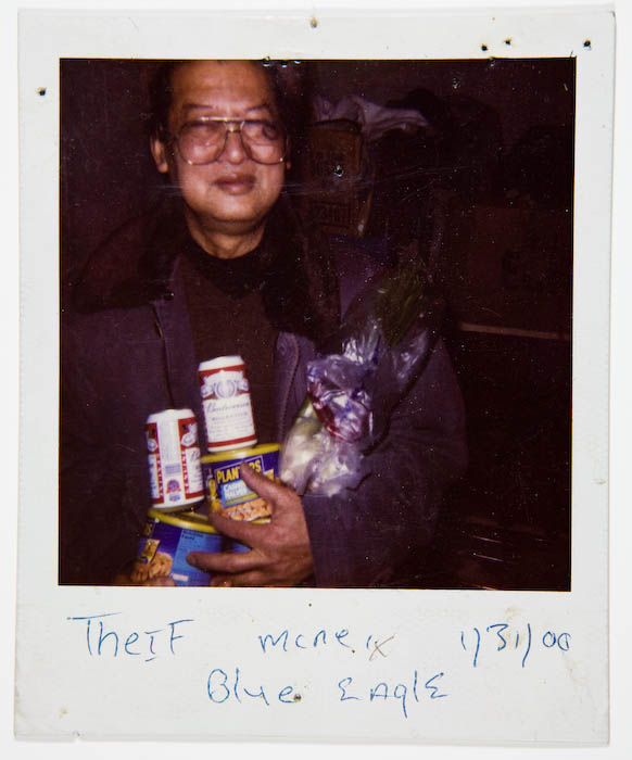 Man in eyeglasses and a dark overcoat holding a Budweiser can, a can of Planters peanuts, another canned item and a bagged bunch of greens against his chest.