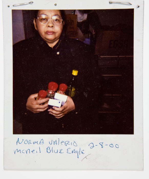 Woman in eyeglasses and a dark coat holding two red-capped spice jars, a bottle of olive oil and a small white box, with egg crates stacked behind her.