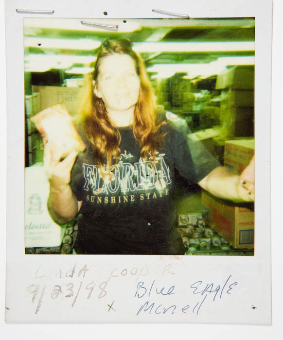 Woman in a Florida Sunshine State T-shirt holding up a wedge of cheese in a crowded stockroom aisle.