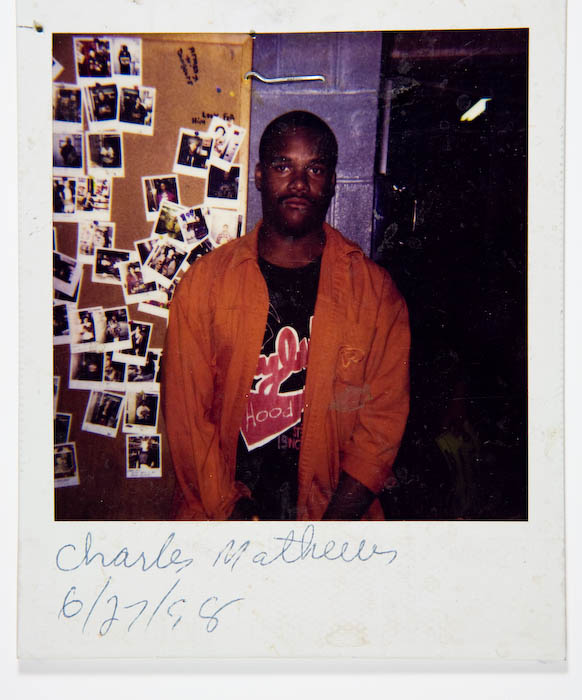 Man in an orange jacket standing beside a corkboard covered with dozens of pinned polaroids of other shoplifters.
