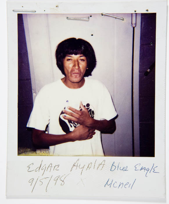 Man in a white graphic T-shirt clutching a small dark package against his chest in front of a white door.