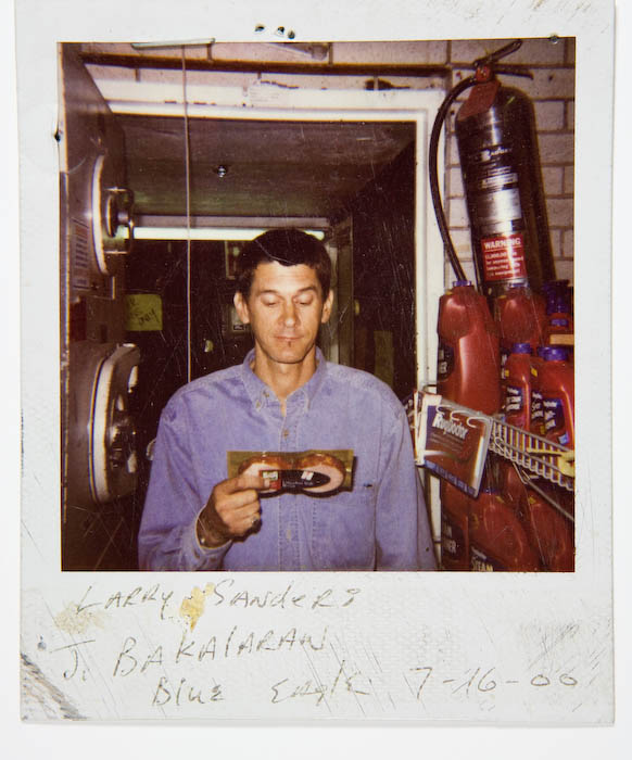 Man in a blue denim shirt looking down at a packaged item in his hands, standing in a stockroom beside shelves of motor oil bottles and a fire extinguisher.