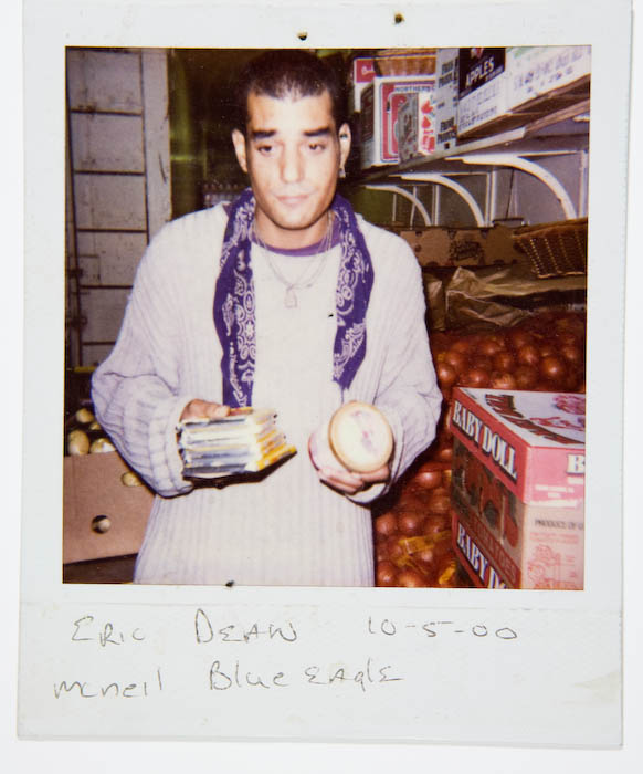 Young man in a cream sweater and blue bandana holding a stack of packaged batteries in one hand and a small round wheel of cheese in the other, beside crates of tomatoes in a stockroom.
