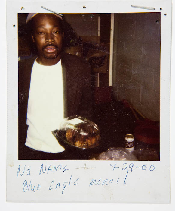 Man in a dark blazer and white T-shirt standing behind a plastic clamshell container of rotisserie chicken on a table.