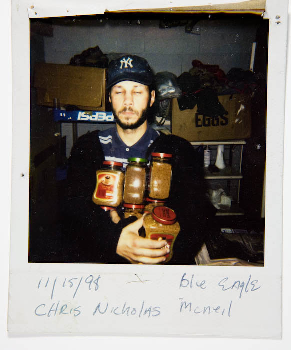 Man in a New York Yankees cap holding four jars of instant coffee and sauce, with cardboard egg and Pepsi crates behind him.