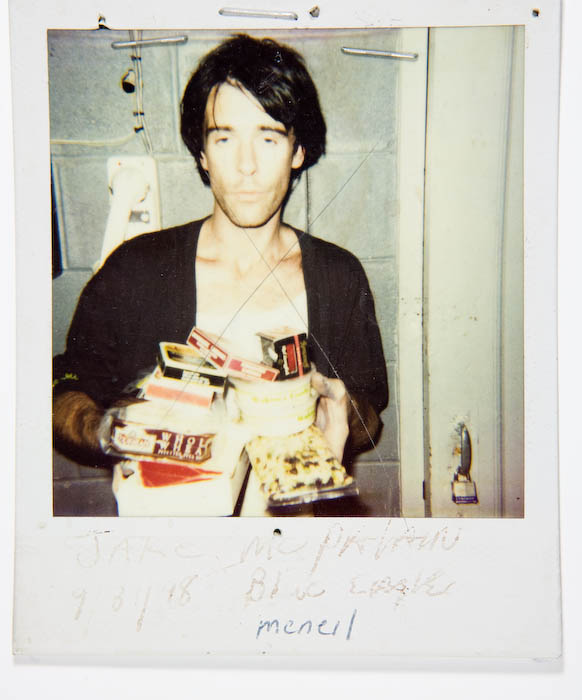 Young man in an open dark shirt holding an armful of boxed goods and a bag of snacks against his bare chest.