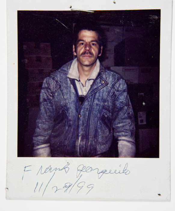 Man in an acid-wash denim jacket standing in front of stacked cardboard cartons in a stockroom.