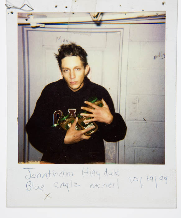 Young man in a dark varsity sweatshirt cradling three jars of instant coffee, standing outside a restroom door marked Men.