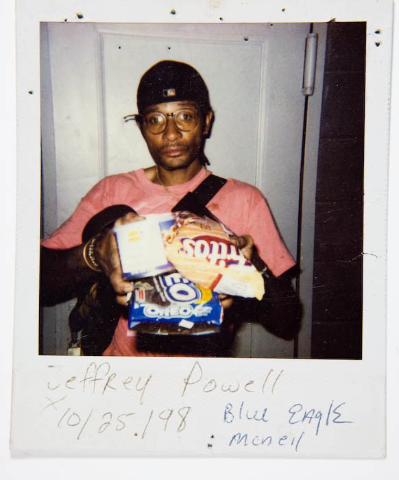 Man in a pink T-shirt, cap and eyeglasses holding bags of Tostitos, Oreo cookies and other snacks.