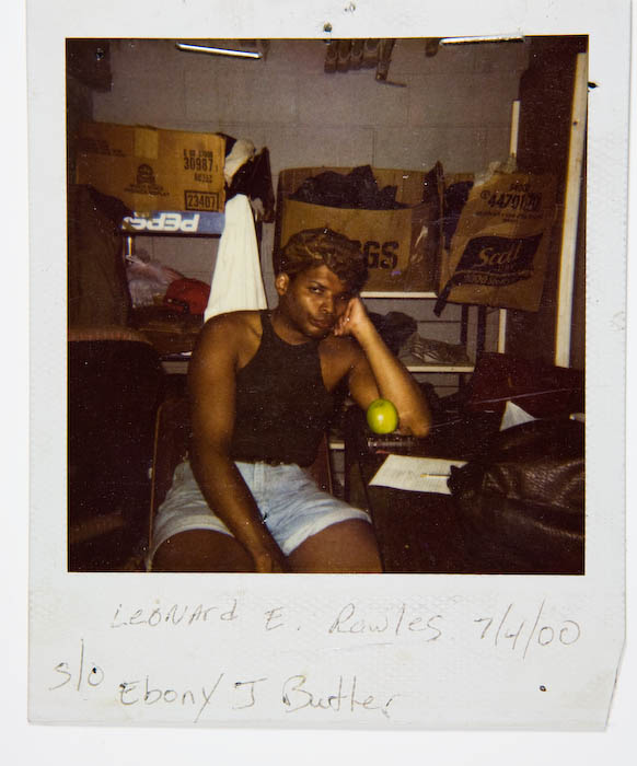 Person in a black tank top and denim shorts seated in a stockroom, head resting on one hand, a green apple on the table beside them.