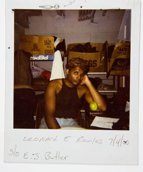 Person in a black tank top seated at a stockroom table with a green apple and paperwork, grocery sacks and crates stacked behind them.