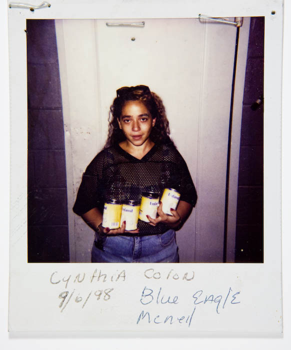 Woman with curly hair in a black mesh jersey and jeans holding four cans of Enfamil infant formula.