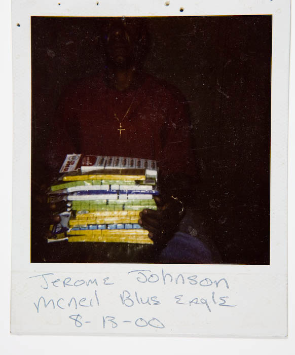 Man in a dark red sweatshirt and cross necklace, lit dimly, holding a tall stack of cigarette cartons.