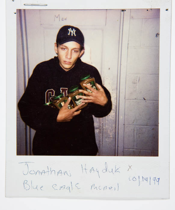 Young man in a New York Yankees cap and black sweatshirt holding three jars of instant coffee, standing outside a restroom door marked Men.