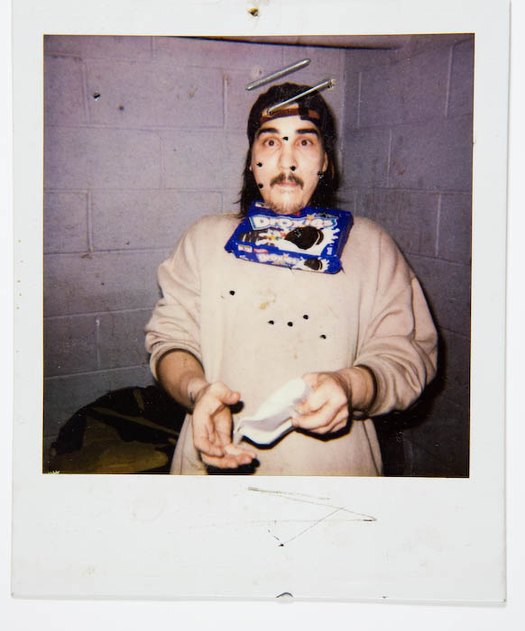 Man in a headband and pale sweatshirt with an opened package of Oreo cookies tucked into his collar, holding out an emptied cookie sleeve against a cinderblock wall.