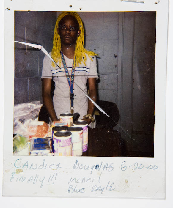 Person with long yellow-blond braids and a lanyard standing behind five tubs of ice cream stacked on a table in a back room.