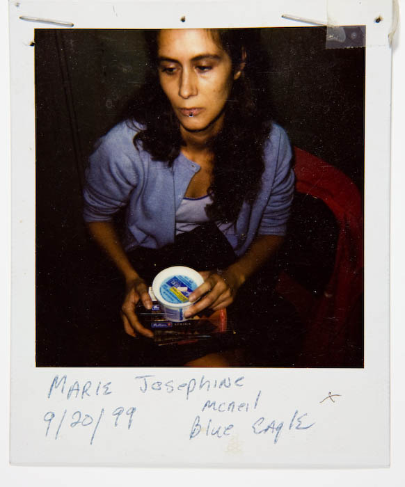 Woman in a pale blue cardigan seated with a plastic tub of food balanced on a stack of boxed hair-color kits in her lap.